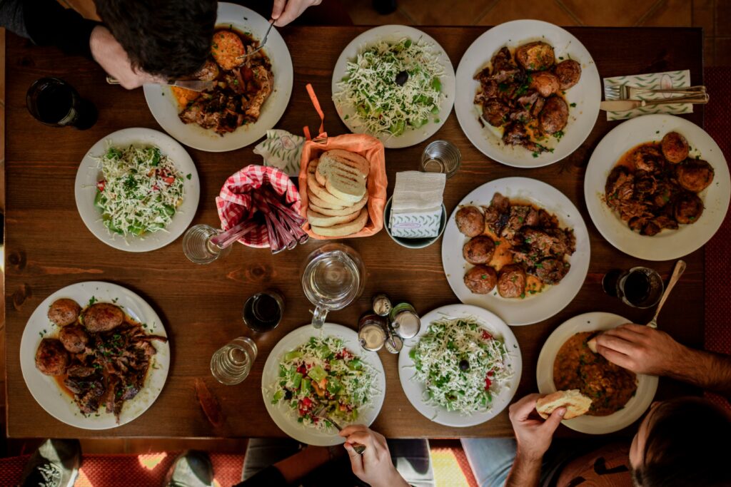 Photo above table crowded with plates of food and seeing hands reach in. Photo by Stefan Vladimirov via unsplash.